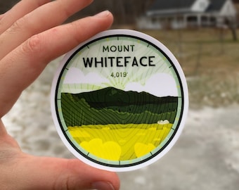 May include: A round sticker featuring a graphic of Mount Whiteface with the text "Mount Whiteface 4,019'". The design includes green and yellow elements, with a white border. The sticker is held in front of a blurred outdoor scene.