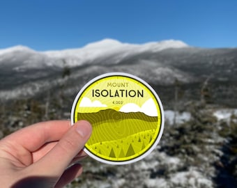 May include: A yellow and white sticker with the text "Mount Isolation 4,003'" and a stylised mountain range design. The sticker is being held in front of a snowy mountain range.