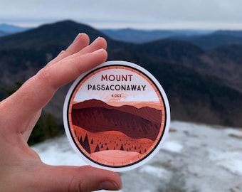 May include: A round sticker featuring a mountain landscape design with the text "Mount Passaconaway 4,043". The sticker has a white border and a color palette of brown, orange, and white. The background shows a mountain range under a cloudy sky.