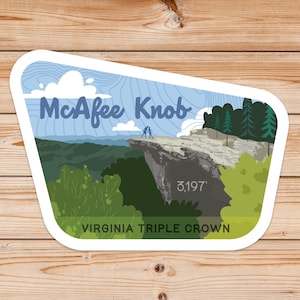 May include: A white sticker with a blue and green illustration of McAfee Knob, a mountain in Virginia. The sticker features the text "McAfee Knob" and "Virginia Triple Crown" and the elevation "3,197'" in white text.