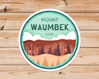 May include: A teal blue and white circular sticker with the text "MOUNT WAUMBEK 4,006'" in a dark green font. The sticker features a stylised illustration of a mountain landscape with brown hills, a white snowy peak, and a forest of dark green trees.
