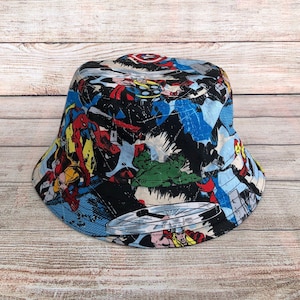 May include: A bucket hat featuring a colorful comic book design with various superheroes. The hat has a black background with blue and white accents, and the brim is lined with a light blue fabric. The hat is made of fabric and is suitable for casual wear.