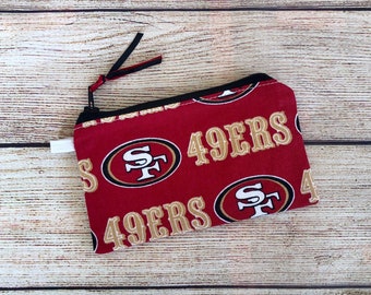 May include: Red zippered pouch with a repeating pattern of the San Francisco 49ers logo and the words "49ers" in gold lettering.