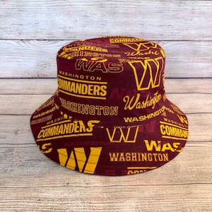 May include: A burgundy bucket hat featuring a repeating pattern of the words "Washington Commanders" and "WAS" in yellow. The hat has a wide brim and a cylindrical crown, ideal for sun protection.