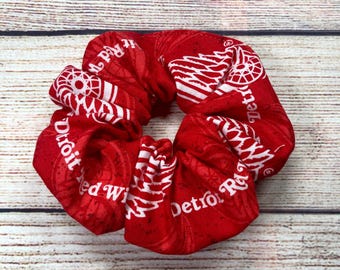 May include: Red scrunchie with white text reading "Detroit Red Wings". The hair accessory is made from patterned fabric. The scrunchie is a hair accessory, suitable for tying up hair.
