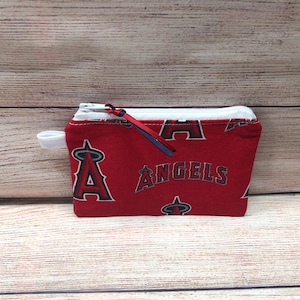Könnte beinhalten: Rote Reißverschlusstasche mit weißem Reißverschluss und einer kleinen weißen Schlaufe an der Seite. Die Tasche ist mit dem Wort "ANGELS" und dem Teamlogo in Weiß gemustert. Ein rotes und blaues Band ist am Reißverschluss befestigt.