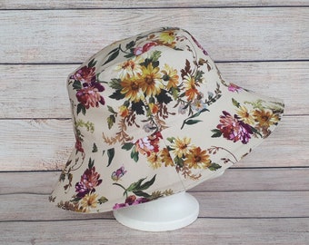 May include: A beige bucket hat with a floral print featuring pink, yellow, and green flowers. The hat has a wide brim and is made of a soft, lightweight fabric.