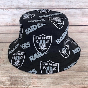 May include: Black bucket hat with a repeating pattern of the Raiders logo and the word "RAIDERS" in white. The hat's design includes the team's logo and the word "RAIDERS" in a bold, stylized font. The hat is made of a durable fabric.