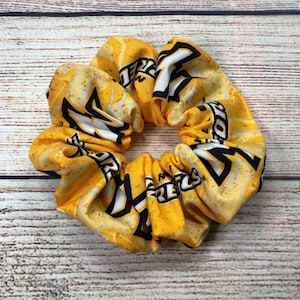 May include: A yellow scrunchie with a repeating pattern of black and white text and shapes. The scrunchie is made of fabric and is designed to hold hair in place. The background is a light brown wooden surface.