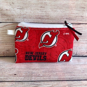 May include: Red zippered pouch with a white zipper and a black pull tab. The pouch is decorated with a repeating pattern of the New Jersey Devils NHL hockey team logo. The text "NEW JERSEY DEVILS" is printed on the pouch.