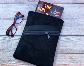 May include: A black suede book sleeve with a zipper closure and a black leather accent. The book sleeve holds a copy of "The Girl in the Spider's Web" by Lisbeth Salander. Tortoiseshell eyeglasses rest on a wooden surface.