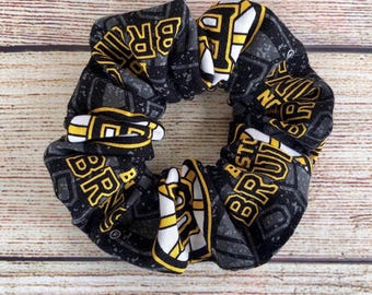 May include: A black and grey scrunchie with yellow and white details. The scrunchie displays the text "BRUINS" in yellow block letters. The fabric has a repeating pattern of the team's logo and name.