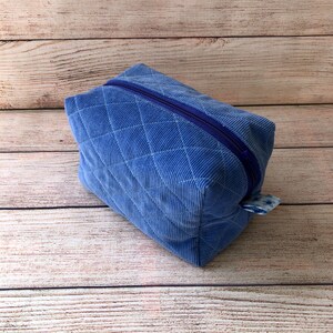 Peut inclure: Une pochette zippée matelassée bleue avec un tirette de fermeture éclair à carreaux blancs et bleus. La pochette est faite d'un tissu doux et texturé.