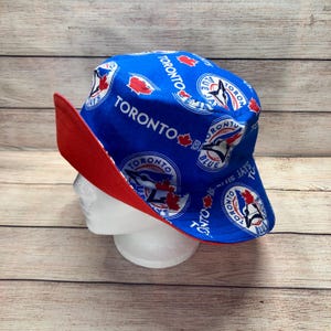 May include: A blue bucket hat with a red brim. The hat features the words "TORONTO BLUE JAYS" and the team logo. The hat is displayed on a white mannequin head against a wooden background.