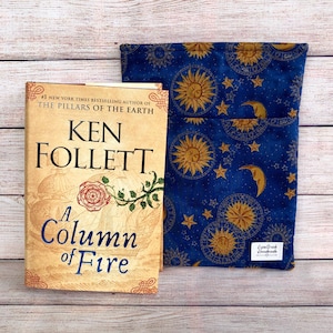 May include: A book and a blue fabric book sleeve. The book is titled "A Column of Fire" by Ken Follett. The book sleeve has a celestial pattern with suns, moons, and stars. The background is a light wood grain.