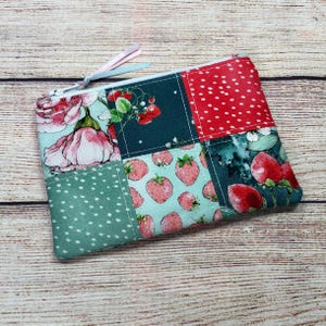 May include: A patchwork zippered pouch featuring floral and fruit patterns. The design includes sections with pink roses, red strawberries, and white polka dots on a teal background. A light blue and pink ribbon is attached to the zipper.
