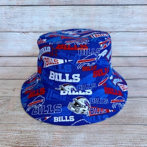 Könnte beinhalten: Ein blauer Fischerhut mit einem durchgehenden Aufdruck des Buffalo Bills Logos, dem Wort Bills in weißen Blockbuchstaben sowie rot-weißen Wimpeldesigns. Das Muster enthält zudem Grafiken von weißen Footballhelmen auf dem Stoff.