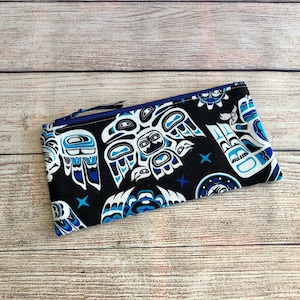 Könnte beinhalten: Eine rechteckige schwarze Reißverschlusstasche mit blauem Reißverschluss und Besatz. Die Tasche hat ein sich wiederholendes Muster aus stilisierten weißen und blauen Designs im indianischen Stil, darunter Vögel und geometrische Formen. Der Hintergrund ist schwarz.