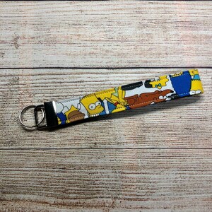 Könnte beinhalten: Ein Schlüsselanhänger aus Stoff mit einem farbenfrohen Cartoon-Design mit gelben, blauen und braunen Figuren. Der Schlüsselanhänger hat einen silbernen Metallring und einen passenden silbernen Metallverschluss. Der Hintergrund ist eine hellbraune Holzoberfläche.