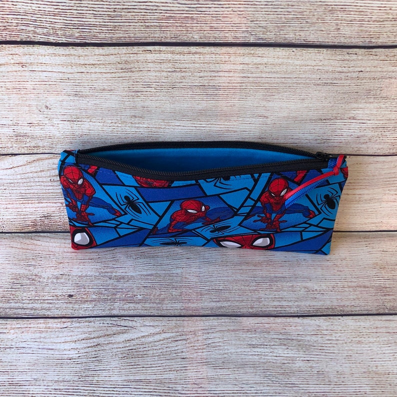 Puede incluir: Una bolsa azul con cremallera con un patr&oacute;n repetido de personajes de Spiderman y telara&ntilde;as. La bolsa est&aacute; abierta y el forro interior es azul.