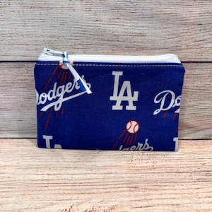 Könnte beinhalten: Eine blaue Münzbörse mit weißem Reißverschluss und Zuglasche. Die Börse ist mit dem Logo der Los Angeles Dodgers und Baseballs in Weiß und Rot gemustert. Der Hintergrund ist ein helles Holzmaserungsmuster.