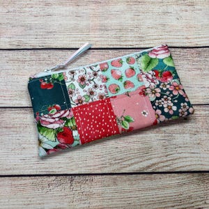 May include: A patchwork zipped pouch with floral and fruit patterns. The design includes strawberries, roses, and other flowers in red, pink, and teal. The pouch has a silver zip and a light-coloured pull.