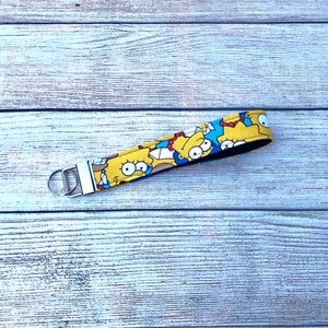 May include: A yellow key fob with a repeating pattern of a cartoon character with blue hair and a yellow dress. The key fob has a silver metal clasp and a black backing. The background is a light wood grain.