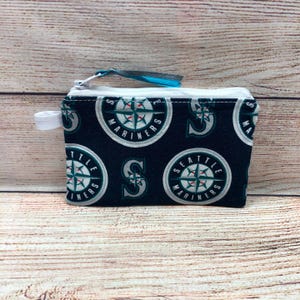 May include: A small, navy blue zippered pouch featuring a repeating pattern of the Seattle Mariners logo in white, teal, and gray. The pouch has a white zipper and a small white loop on one side.