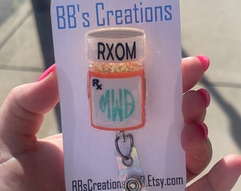 May include: A white card with the text "BB's Creations" at the top and "BBsCreations.Etsy.com" at the bottom. A clear plastic pill bottle with orange glitter and the text "RXOM" is attached to a retractable badge holder.