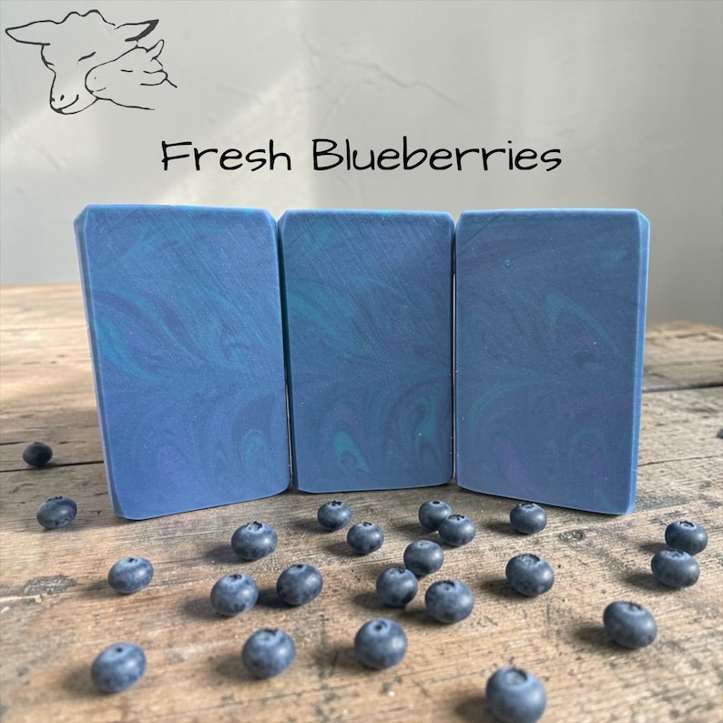 May include: Three rectangular bars of soap with a blue and teal swirl pattern, arranged on a wooden surface. Scattered around the soap are fresh blueberries. The text "Fresh Blueberries" is above the soap.