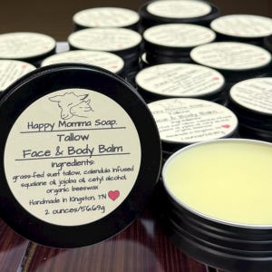 May include: A collection of black metal tins of Happy Momma Soap Face & Body Balm. The labels are white with black text and a sheep illustration. The balm is a light yellow color and the label states the product contains 2 ounces/56.69g.