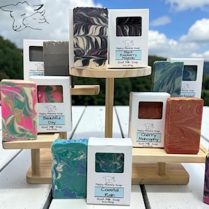 May include: A collection of rectangular goat milk soap bars arranged on a tiered wooden stand. Each bar features unique swirl patterns in colors including pink, green, blue, black, and red. White paper labels display the brand name Happy Momma Soap.