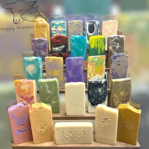 May include: A wooden shelf display with a variety of colorful handmade soap bars. The soap bars are arranged in rows and feature different designs and patterns. The shelf is labeled "Happy Momma Soap".