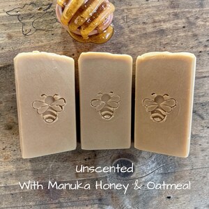 May include: Three rectangular bars of light brown soap, each featuring a stamped bee design in the center. Above the soap sits a wooden honey dipper coated in golden honey. The text below reads: Unscented With Manuka Honey & Oatmeal.