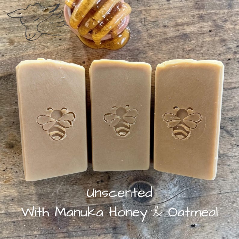 May include: Three rectangular bars of light brown soap, each featuring a stamped bee design in the center. Above the soap sits a wooden honey dipper coated in golden honey. The text below reads: Unscented With Manuka Honey & Oatmeal.