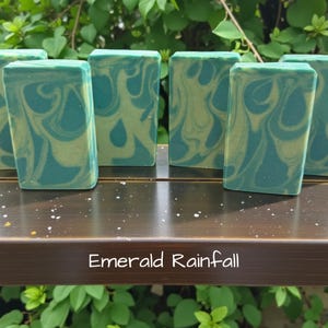 Puede incluir: Pastillas de jab&oacute;n rectangulares con un patr&oacute;n jaspeado en colores verde azulado y crema. Las pastillas est&aacute;n dispuestas en fila y el texto "Emerald Rainfall" est&aacute; impreso en blanco debajo, sobre la superficie marr&oacute;n oscuro en la que descansan.