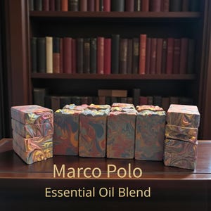 May include: Several bars of handmade soap with a marbled design in shades of red, yellow, blue, and orange. The soap is stacked in groups and arranged on a dark wooden surface. The text "Marco Polo Essential Oil Blend" is displayed below the soap.