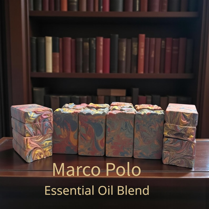 May include: Several bars of handmade soap with a marbled design in shades of red, yellow, blue, and orange. The soap is stacked in groups and arranged on a dark wooden surface. The text "Marco Polo Essential Oil Blend" is displayed below the soap.