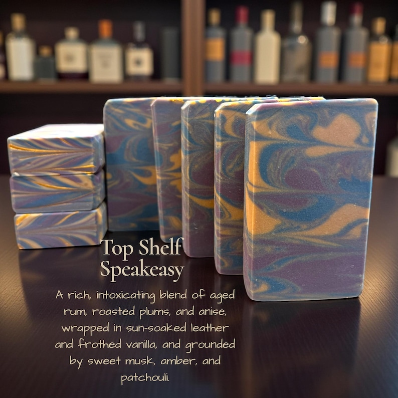 May include: Artisan soap bars with a swirled design in shades of blue, purple, and gold. The bars are rectangular and stacked, with the text "Top Shelf Speakeasy" visible. The soap is described as a blend of aged rum, plums, and anise.
