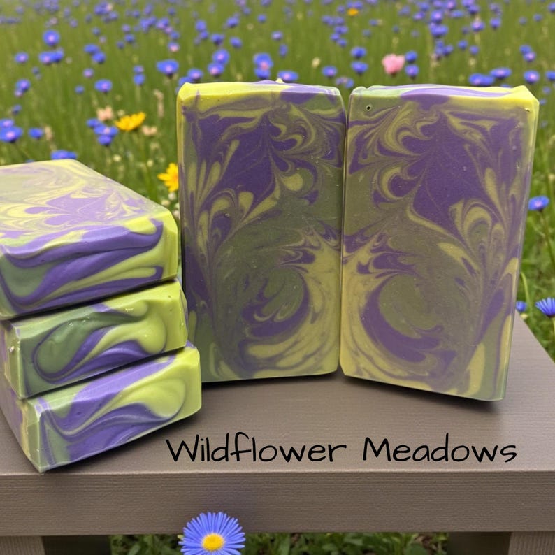 May include: Three rectangular bars of soap with a marbled purple and lime green swirl pattern. The text Wildflower Meadows is printed in black on the surface below the soap bars. The soap has a smooth, solid, and opaque appearance.