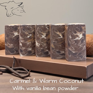 May include: Five rectangular bars of soap with a marbled pattern of dark brown, tan, and white swirls. The soap is displayed with two whole coconuts and dried vanilla beans. The text reads: Carmel & Warm Coconut With vanilla bean powder.