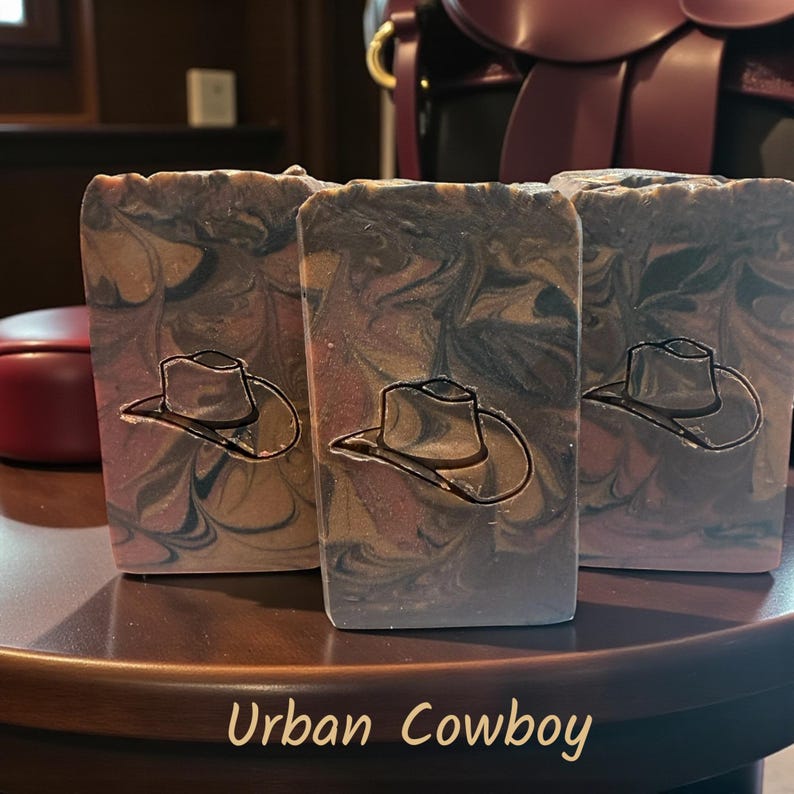 Puede incluir: Tres barras rectangulares de jab&oacute;n con un patr&oacute;n jaspeado en tonos marr&oacute;n, negro y bronceado. Cada barra est&aacute; grabada con el contorno detallado de un sombrero de vaquero. El texto "Urban Cowboy" aparece en una fuente color bronceado claro.