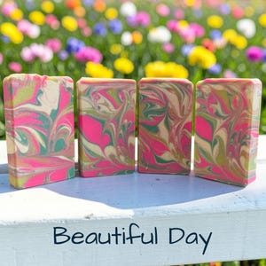 May include: Four rectangular bars of handmade soap with a vibrant swirl pattern of pink, green, and beige. The bars are arranged side by side on a white surface, with a blurred floral background. The text "Beautiful Day" is at the bottom.