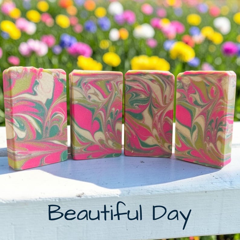 May include: Four rectangular bars of handmade soap with a vibrant swirl pattern of pink, green, and beige. The bars are arranged side by side on a white surface, with a blurred floral background. The text "Beautiful Day" is at the bottom.