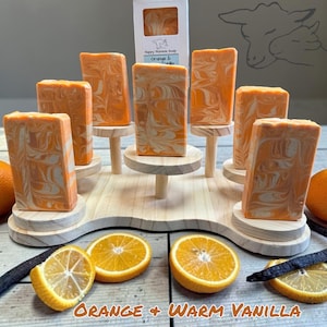 Puede incluir: Barras rectangulares de jab&oacute;n con remolinos naranjas y blancos dispuestas en un soporte de madera escalonado. Una caja blanca con la etiqueta Happy Momma Soap Orange & Vanilla se sit&uacute;a en el centro. Rodajas de naranja y vainas de vainilla en la base.