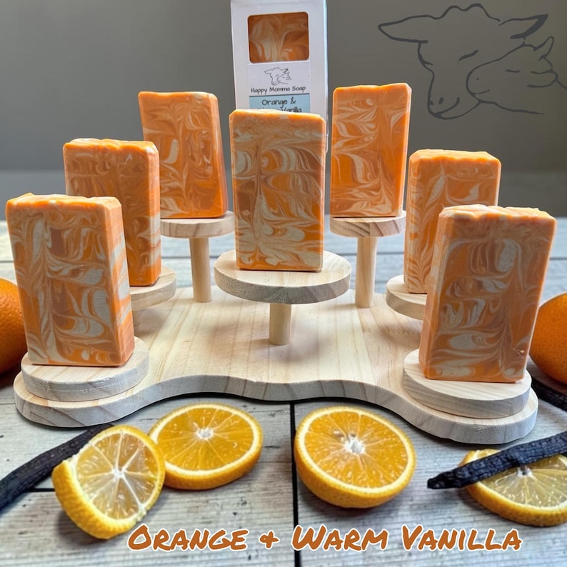 Puede incluir: Barras rectangulares de jab&oacute;n con remolinos naranjas y blancos dispuestas en un soporte de madera escalonado. Una caja blanca con la etiqueta Happy Momma Soap Orange & Vanilla se sit&uacute;a en el centro. Rodajas de naranja y vainas de vainilla en la base.