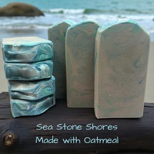 May include: A stack of four rectangular soap bars and two upright soap bars featuring a marbled pattern of teal, blue, and beige tones. The text "Sea Stone Shores Made with Oatmeal" is printed in light blue at the bottom of the image.