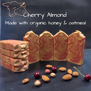 May include: Four rectangular bars of soap with a red and gold marbled pattern, stacked and standing upright. Text reads: Cherry Almond, Made with organic honey & oatmeal. Scattered around are whole almonds and dark red cherries on stems.