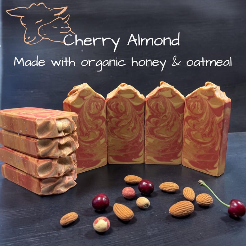 May include: Four rectangular bars of soap with a red and gold marbled pattern, stacked and standing upright. Text reads: Cherry Almond, Made with organic honey & oatmeal. Scattered around are whole almonds and dark red cherries on stems.