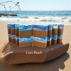 Puede incluir: Una colecci&oacute;n de pastillas de jab&oacute;n rectangulares dispuestas en fila, con capas horizontales marrones y azules con remolinos blancos que recuerdan a las olas del mar. El texto Coco Beach est&aacute; impreso en blanco, junto a un dibujo de dos ovejas.